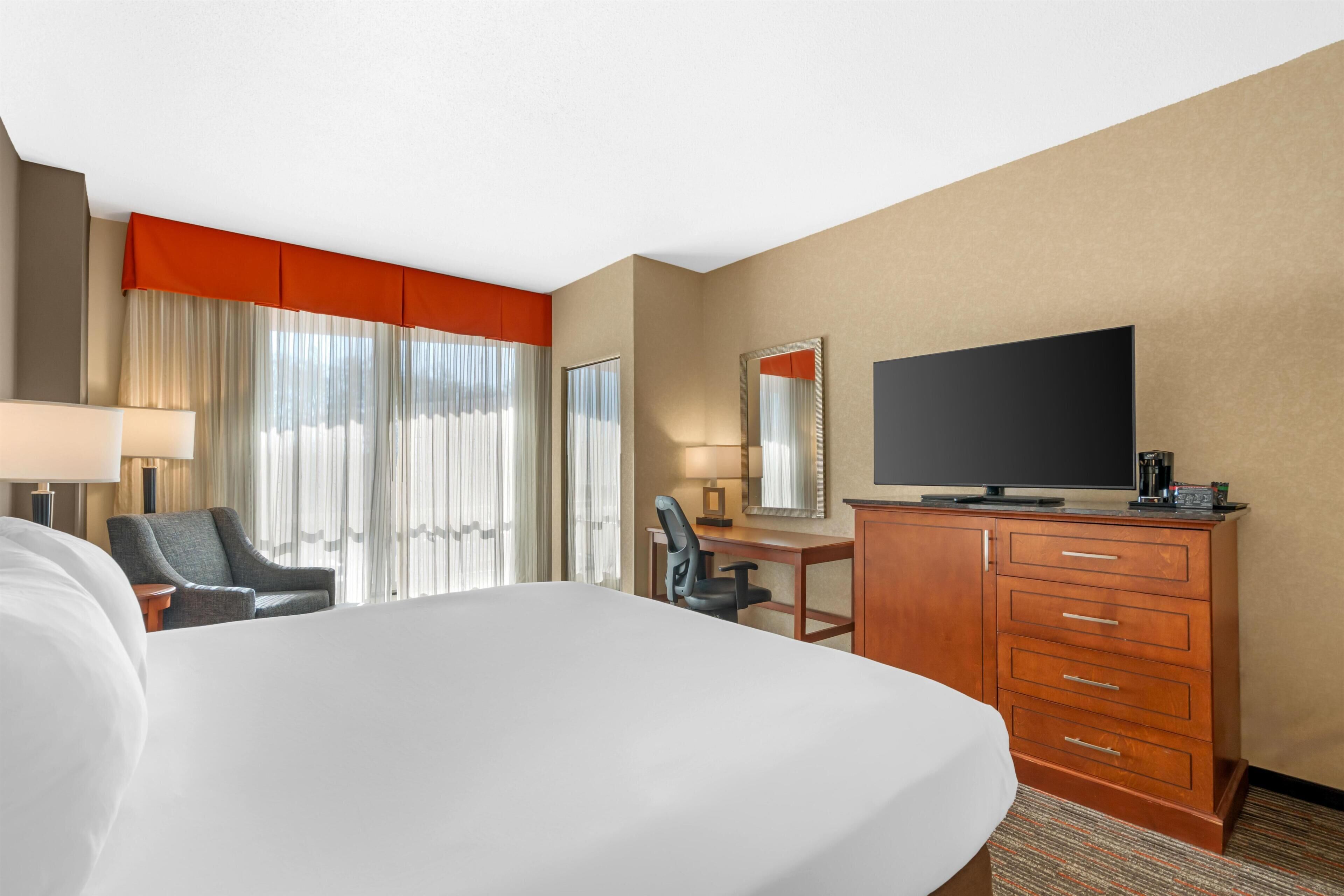 Quarto de hotel com cama, escrivaninha, cadeira, televisão e cômoda.