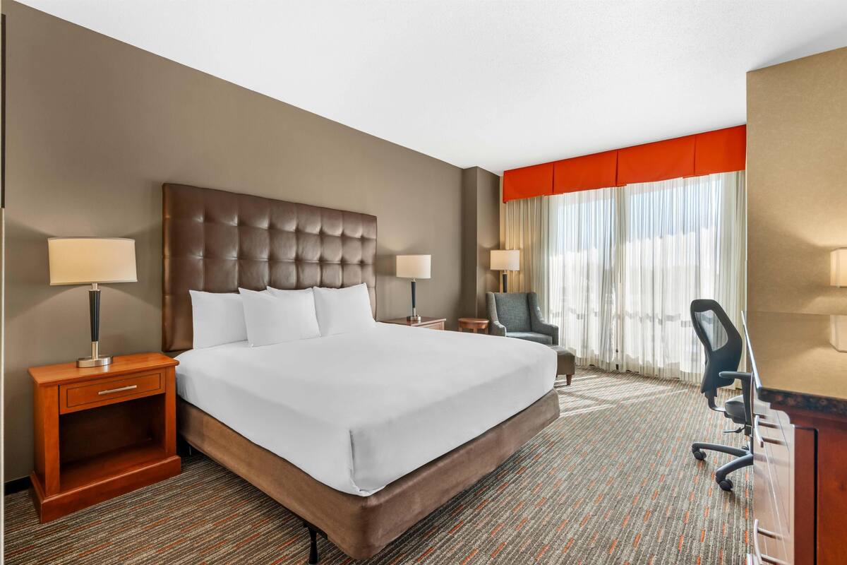 Une chambre d’hôtel avec un grand lit, deux tables de chevet avec des lampes, un bureau avec une chaise et une fenêtre avec des rideaux.