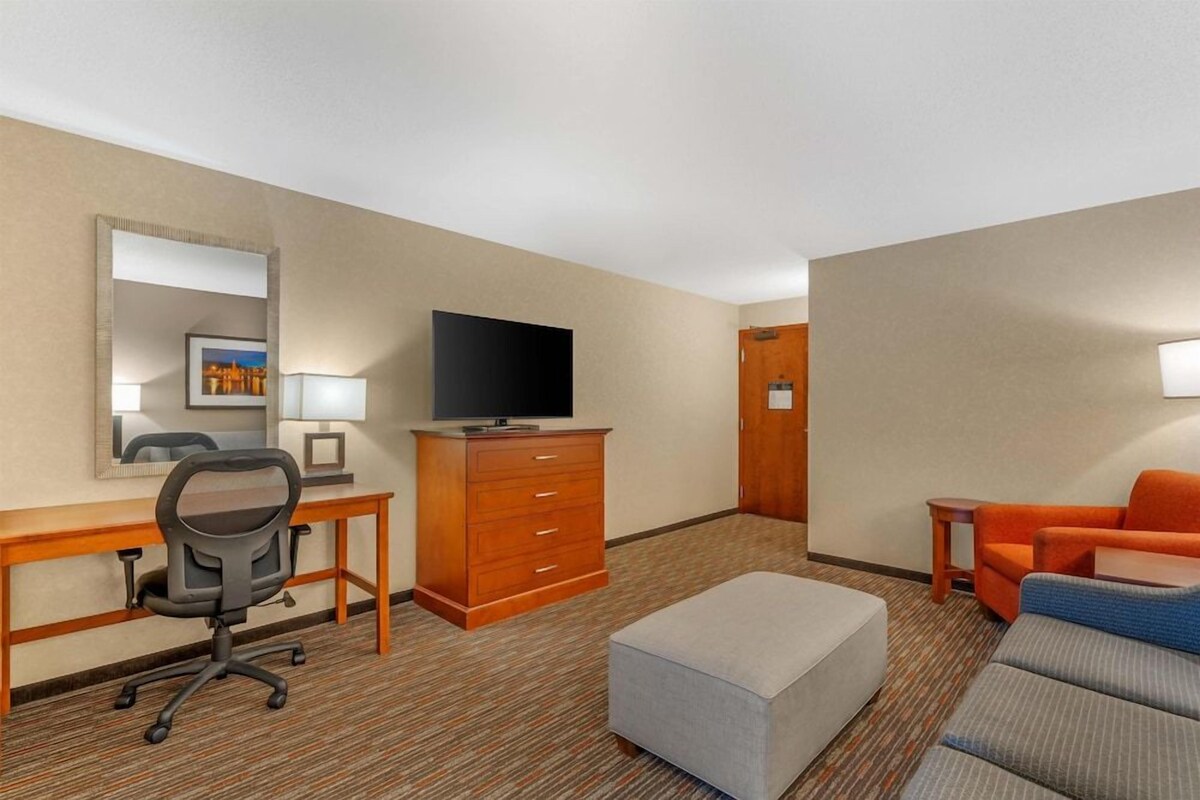 Een hotelkamer met een bureau, stoel, televisie, dressoir, sofa en pouf.