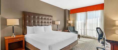 A hotel room with a large bed, two bedside tables with lamps, a desk with a chair, and a window with curtains.