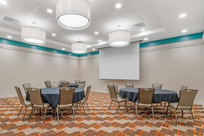Une salle de conférence équipée de tables, de chaises, d’un écran de projection et d’un tapis à motifs.