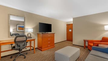 A hotel room with a desk, chair, television, dresser, sofa, ottoman, and a mirror.