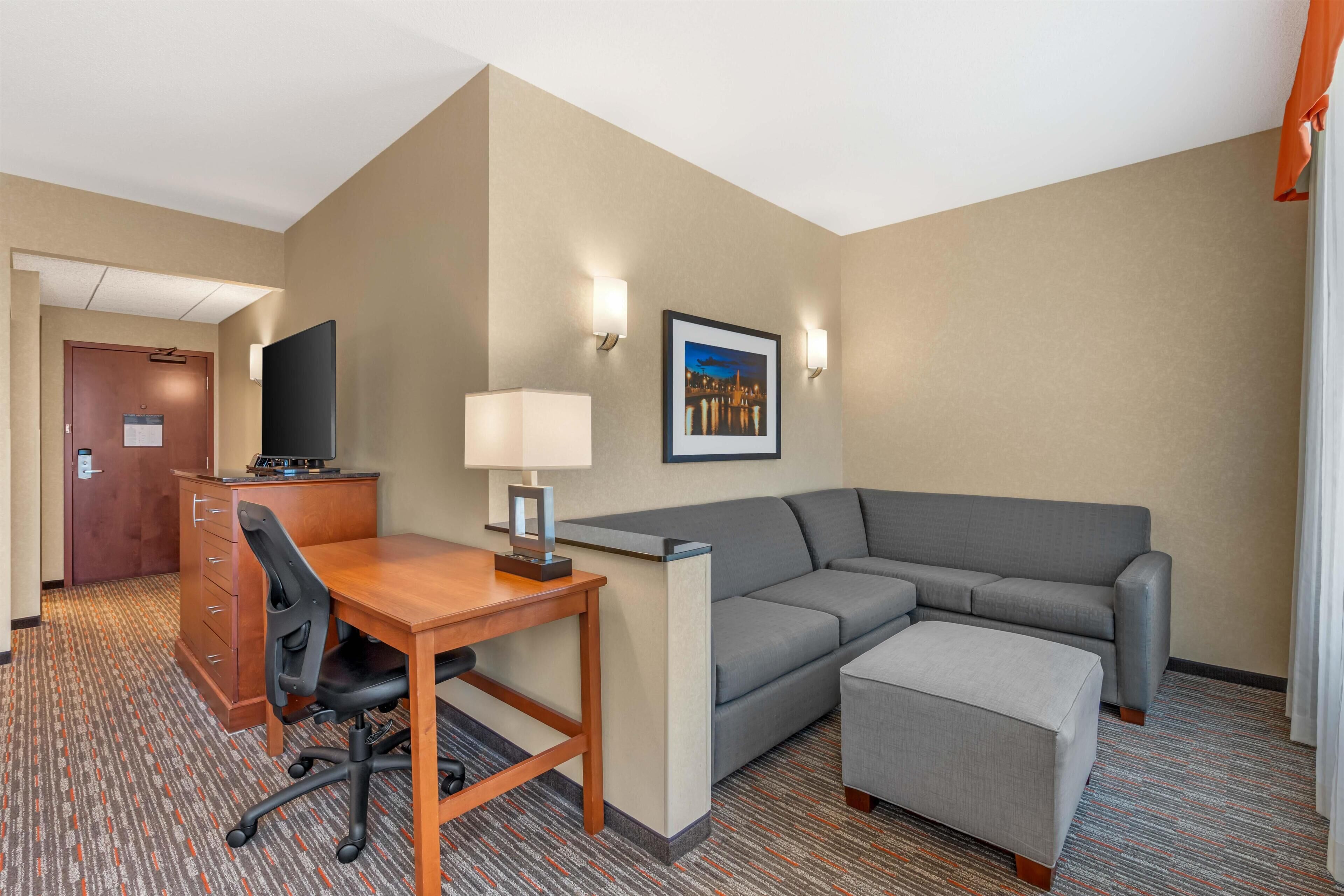A hotel room with a desk, chair, sofa, ottoman, and a television.