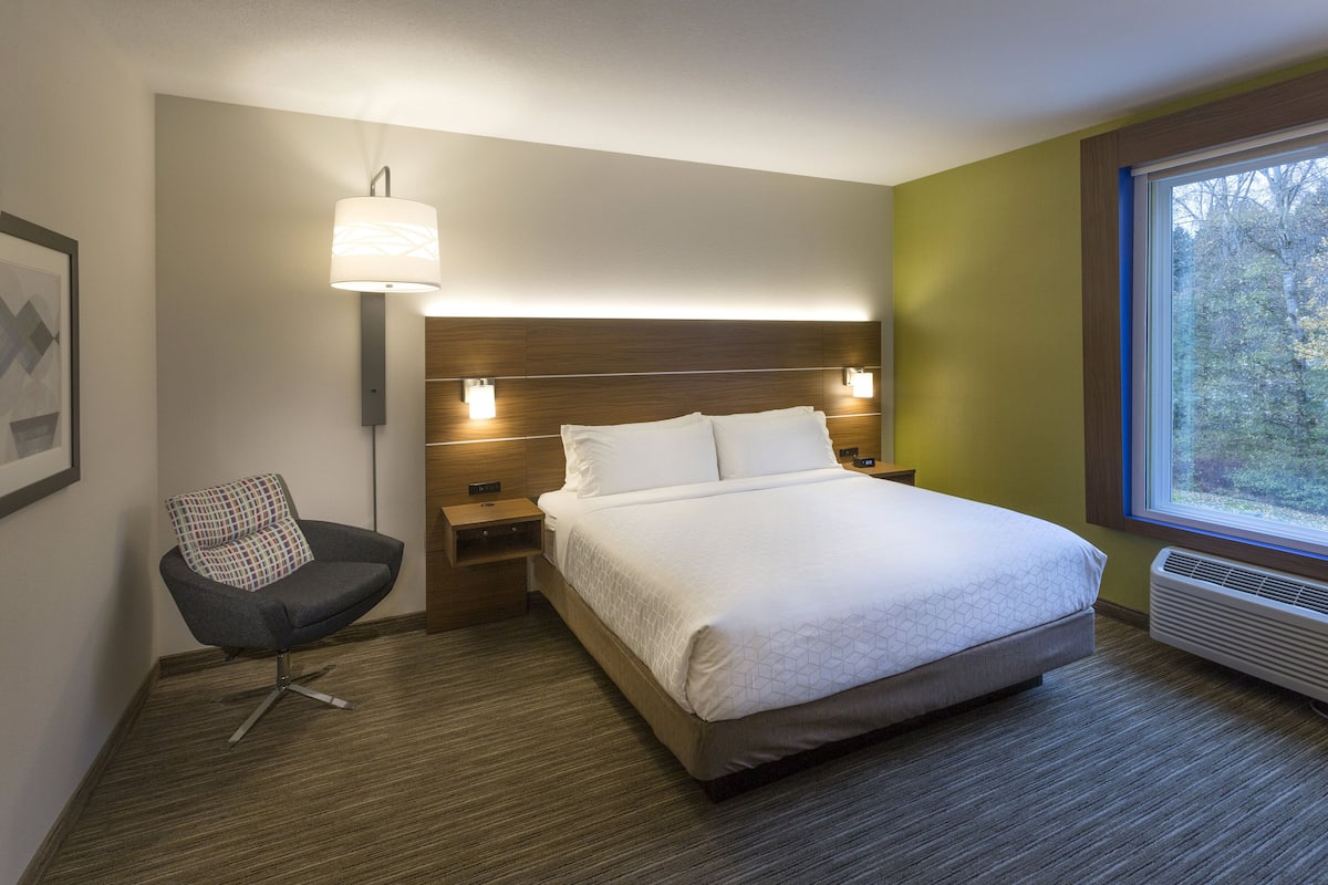 A modern hotel room with a large bed, bedside tables, a chair, a wall-mounted lamp, and a window with a view of trees.