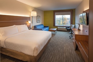 Quarto de hotel com uma cama grande, uma escrivaninha, uma cadeira, um sofá e uma janela ampla com vista para prédios.