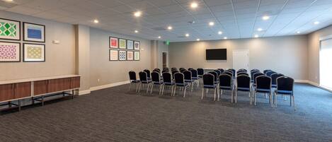A conference room with a carpeted floor, a row of chairs, a wooden cabinet, and framed artwork on the wall.