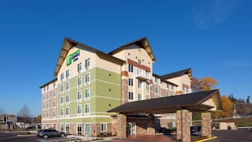 Ein mehrstöckiges Hotel mit einer Fassade in Grün- und Beigetönen, einem überdachten Eingangsbereich und einem Parkplatz mit Autos.