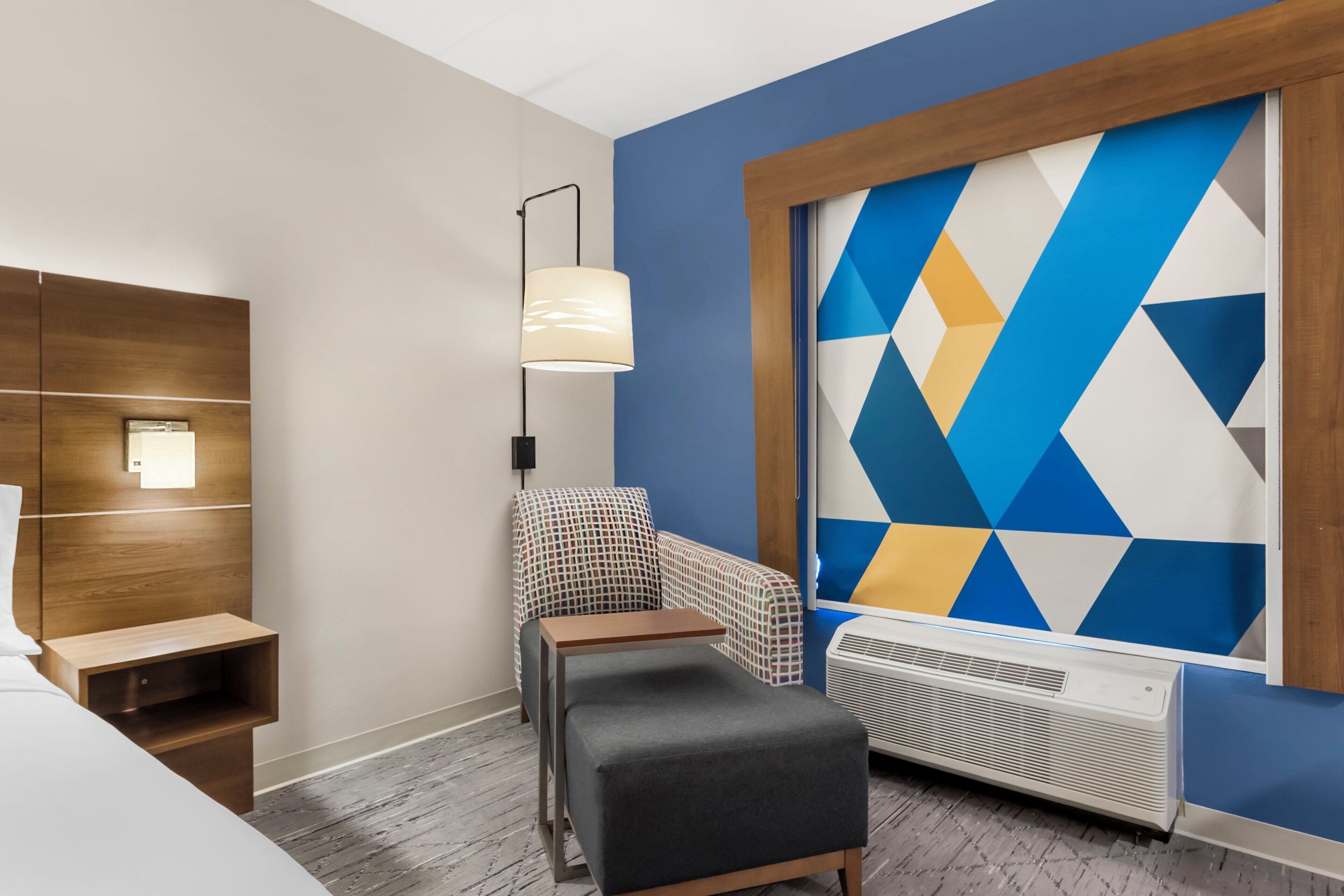 A hotel room with a bed, a nightstand, a chair, a table, a lamp, a wall unit, and a large geometric-patterned wall.