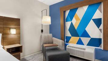 A hotel room with a bed, a nightstand, a chair, a table, a lamp, a wall unit, and a large geometric-patterned wall.