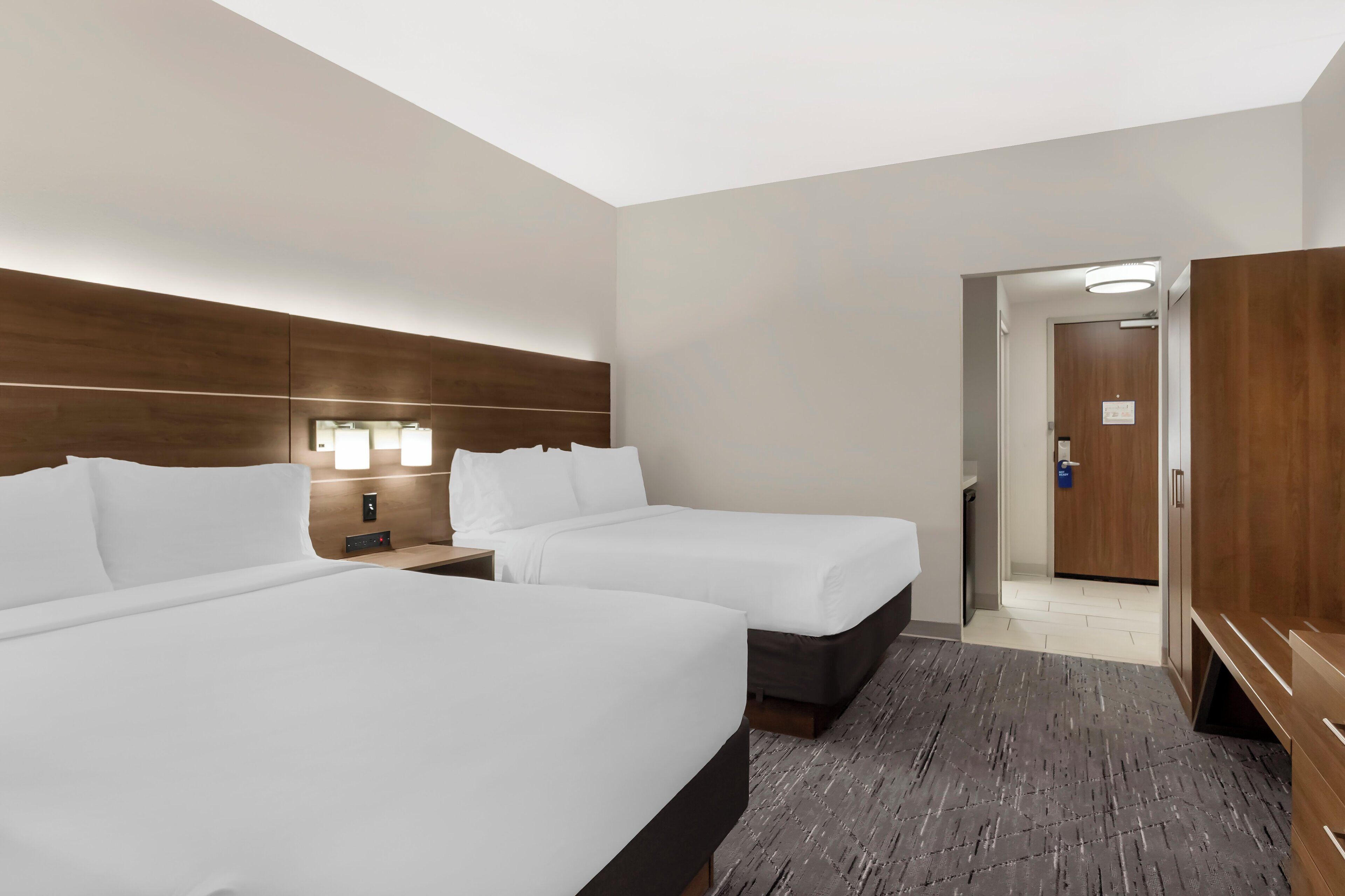 Habitación de hotel con dos camas, cabecero de madera, lámparas de noche, un armario y una puerta que da al baño.