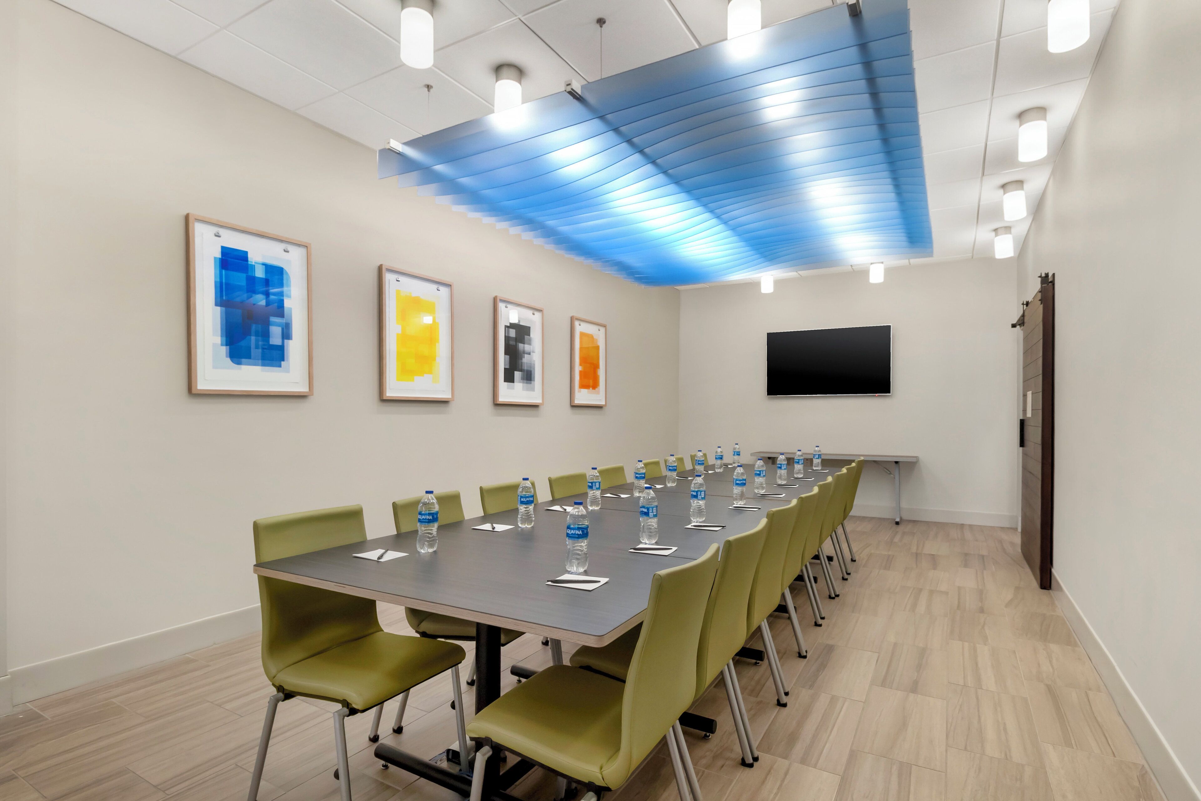 A conference room with a long table, green chairs, framed artworks on the wall, and a flat-screen TV.