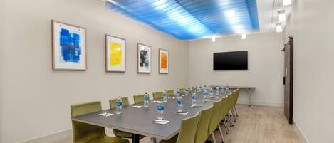 A conference room with a long table, green chairs, framed artworks on the wall, and a flat-screen TV.