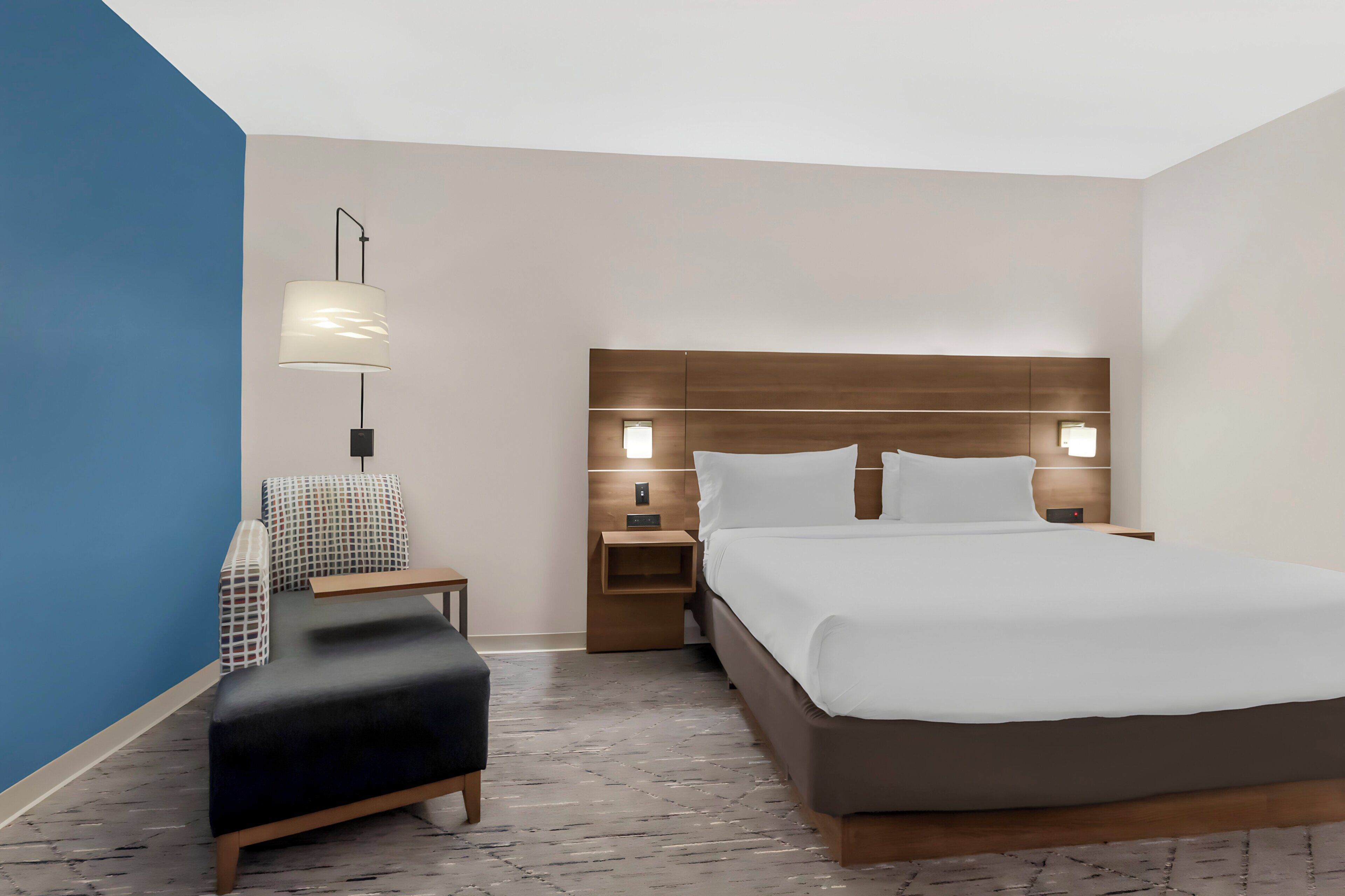 A modern hotel room with a large bed, a desk, a chair, and a nightstand with a lamp.