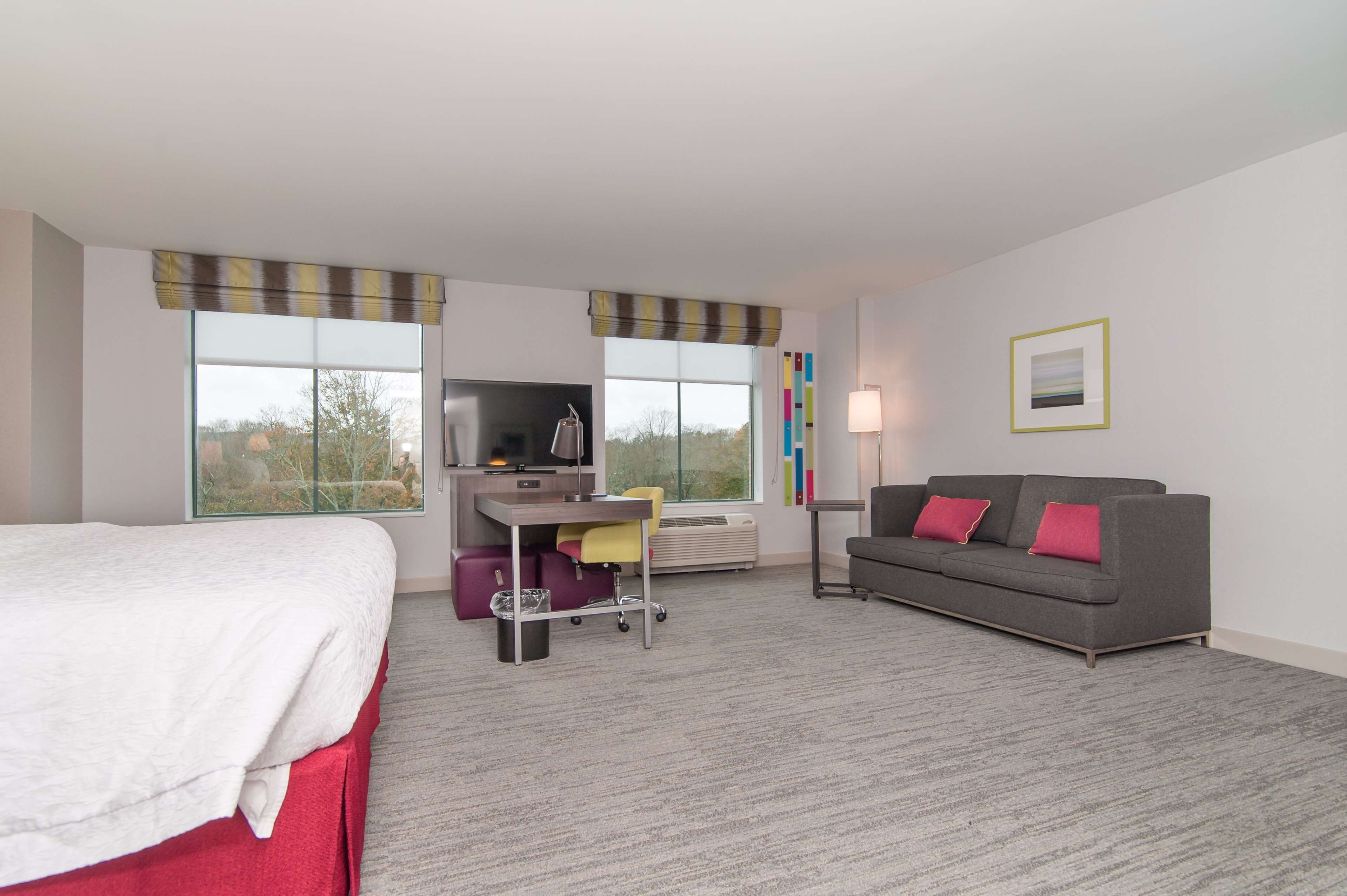 A hotel room with a bed, a desk with a chair, a grey sofa, and a TV.