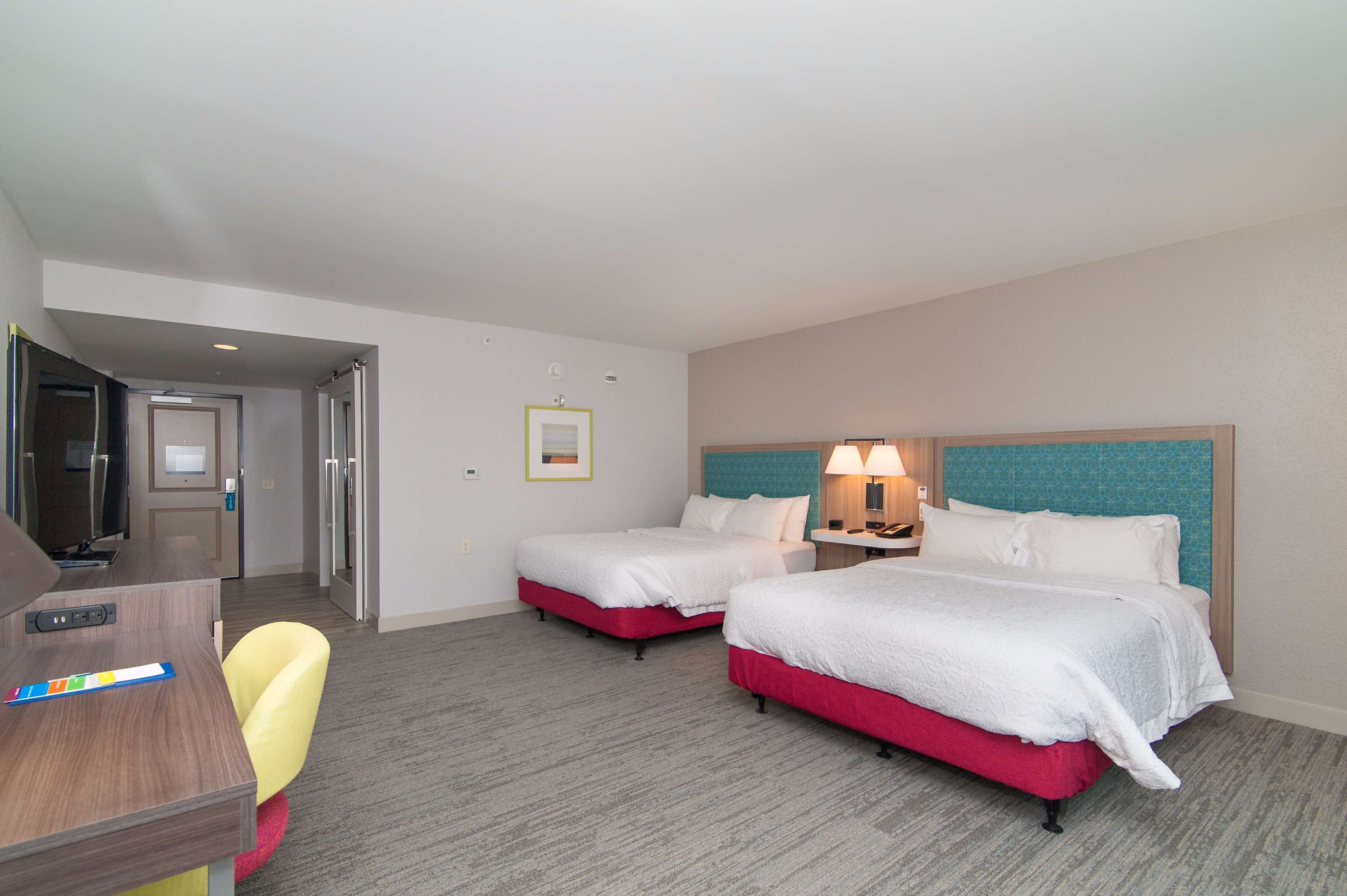 A hotel room with two beds, a desk, a chair, a lamp, and a door leading to a bathroom.