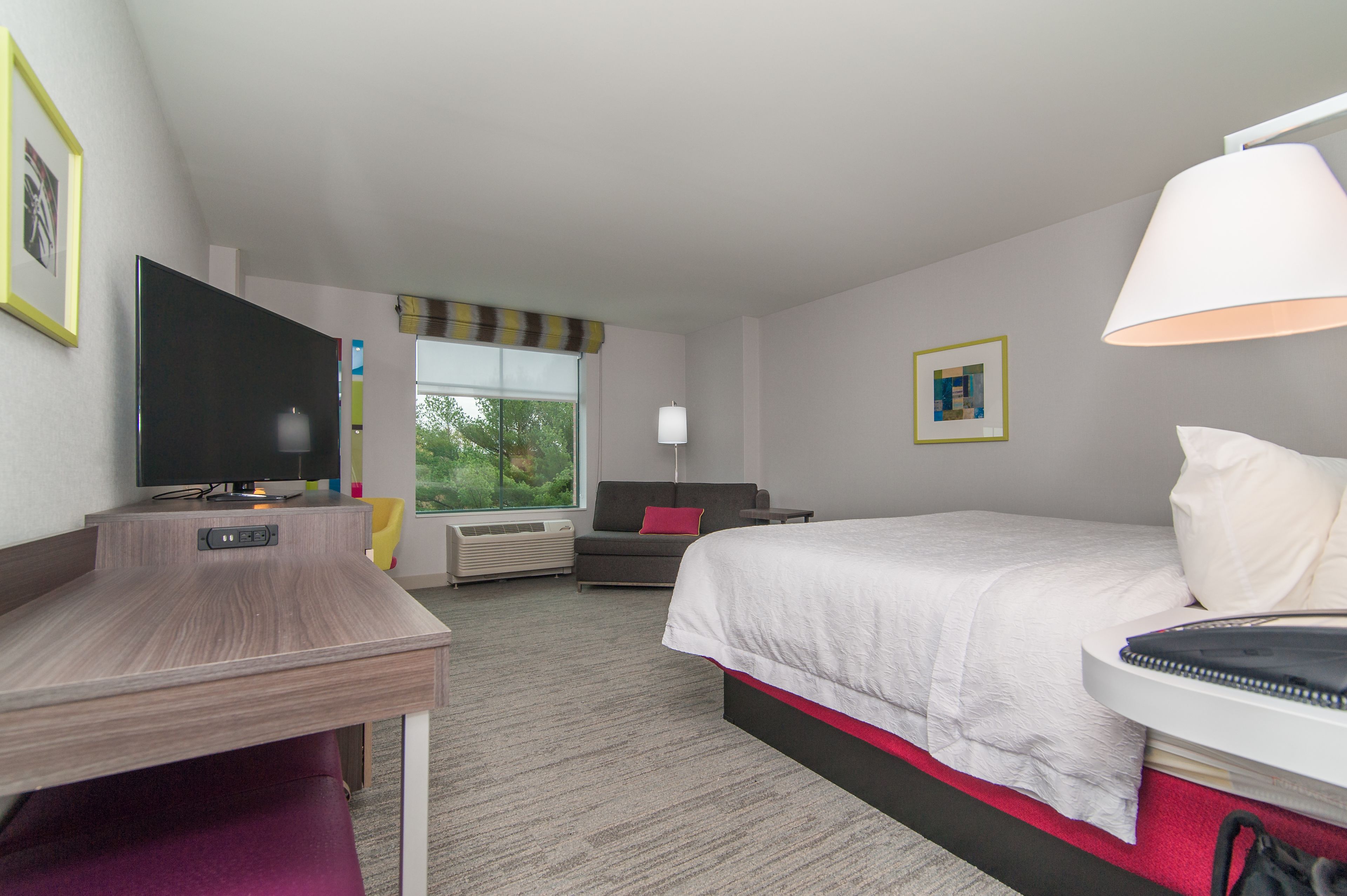 A hotel room with a bed, a desk, a TV, a window, and a lamp.