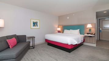 Habitación de hotel con cama, mesita de noche con lámpara, una pequeña mesa, sofá con cojín y un cuadro en la pared.
