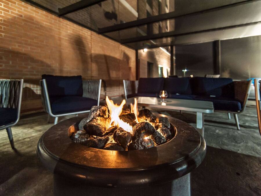 A fire pit with burning logs in a modern outdoor seating area with brick walls and metal chairs.