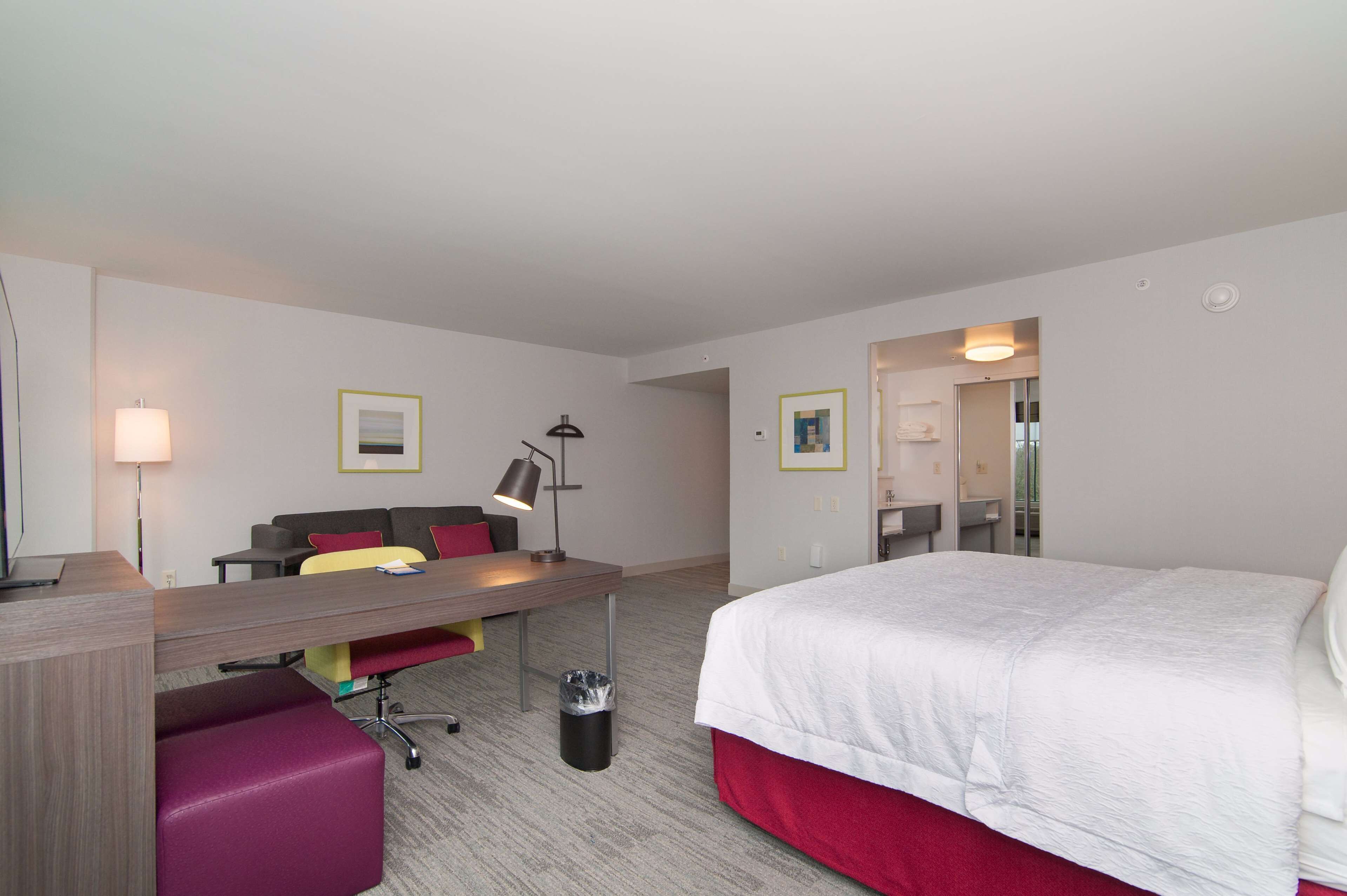 A hotel room with a bed, a desk with a chair, a sofa, a lamp, and a trash can.