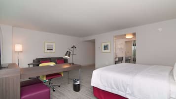 A hotel room with a bed, a desk with a chair, a sofa, a lamp, and a trash can.
