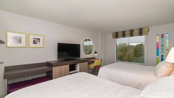 A hotel room with two beds, a desk with a TV, a mirror, and a window with a view of trees.