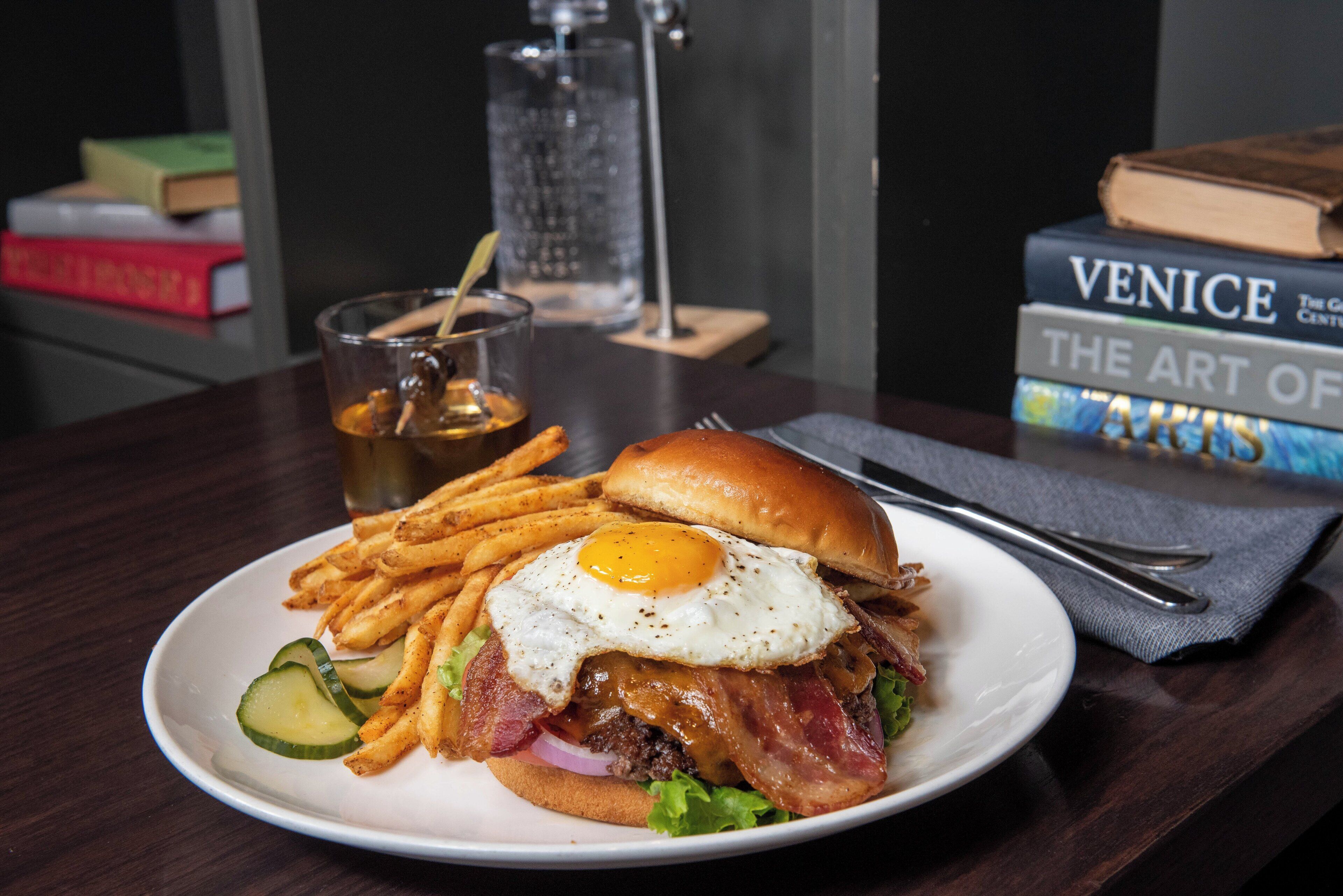 A plate of food with a sandwich, fries, and a drink on a table with books in the background.