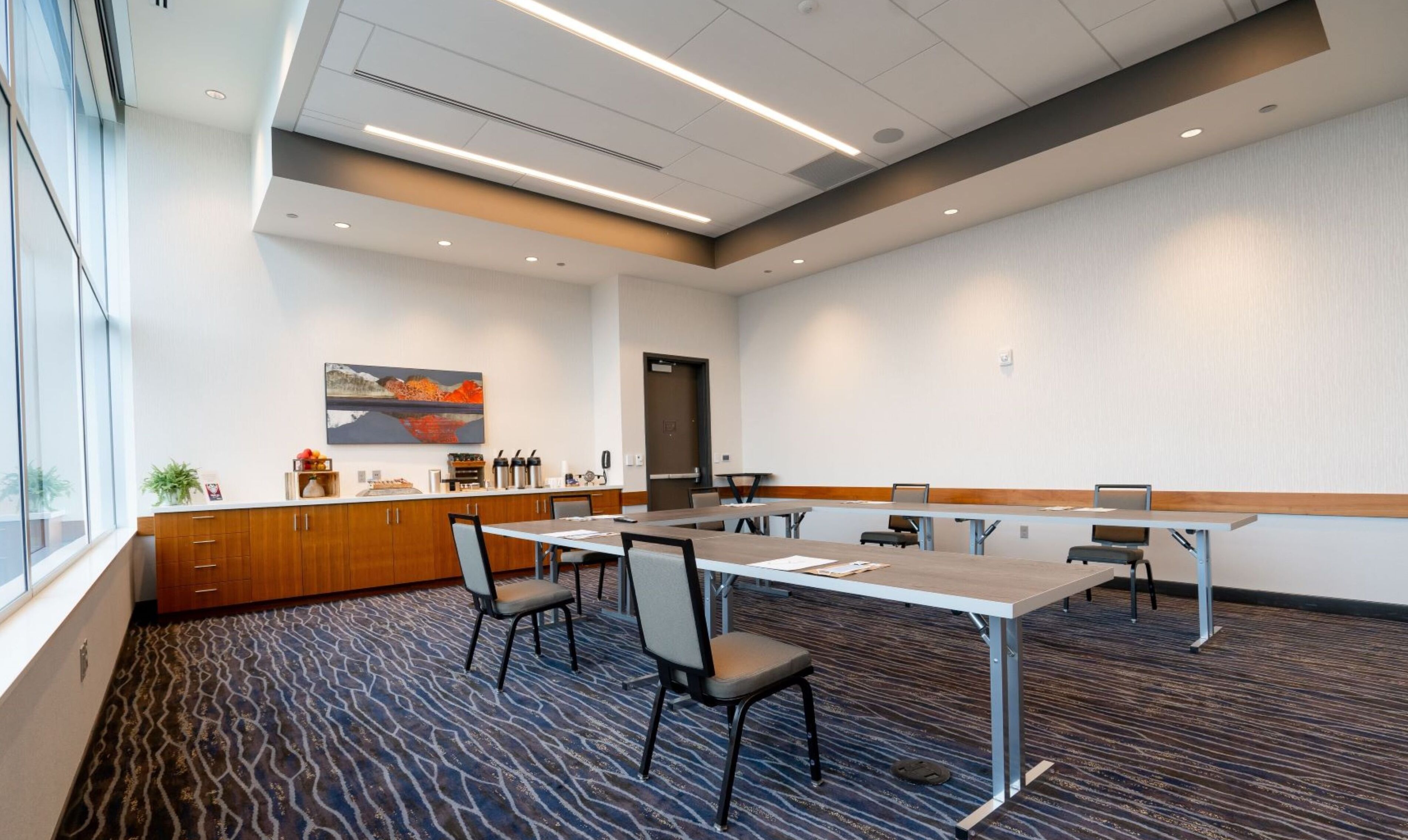 A conference room with a long table, chairs, a painting, and a kitchen area with cabinets and appliances.