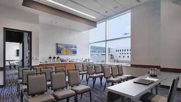 Un salón de conferencias con una larga mesa y varias sillas, una estación de café y un ventanal con vistas a edificios y un avión.
