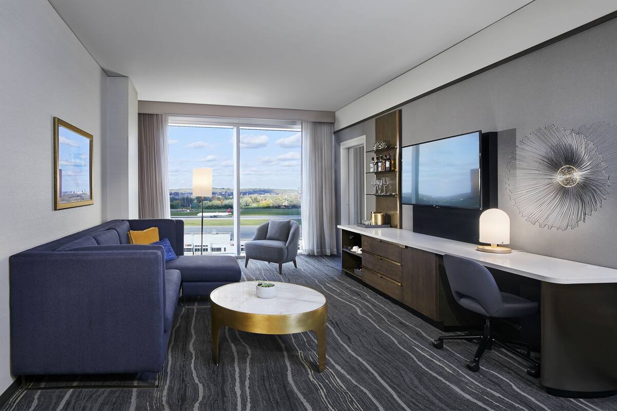 A modern hotel room with a sofa, a desk, a chair, a television, and a large window with a city view.