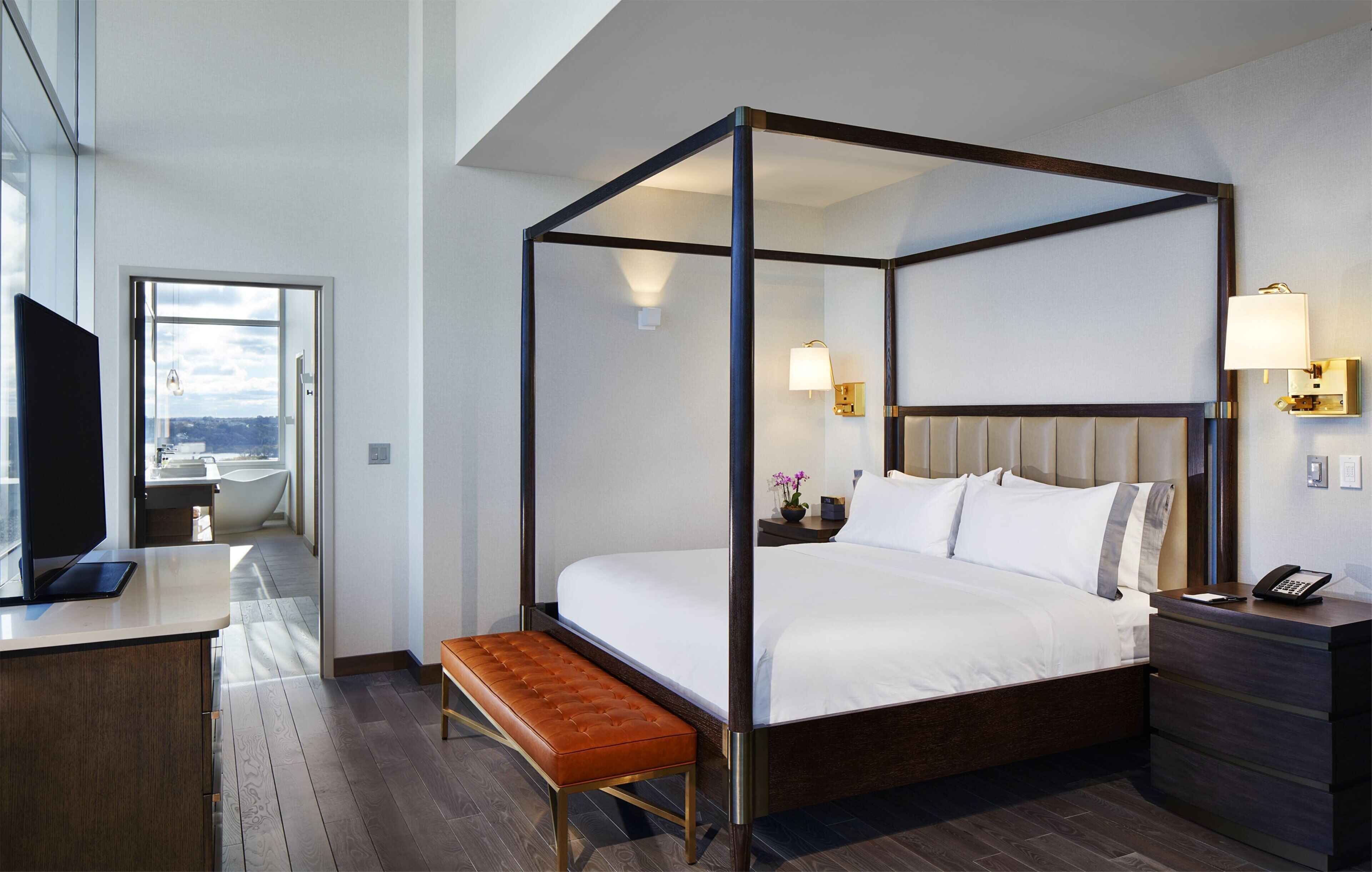 A modern hotel room with a four-poster bed, a wooden bench, a desk with a flat-screen TV, and a view of the city through a large window.