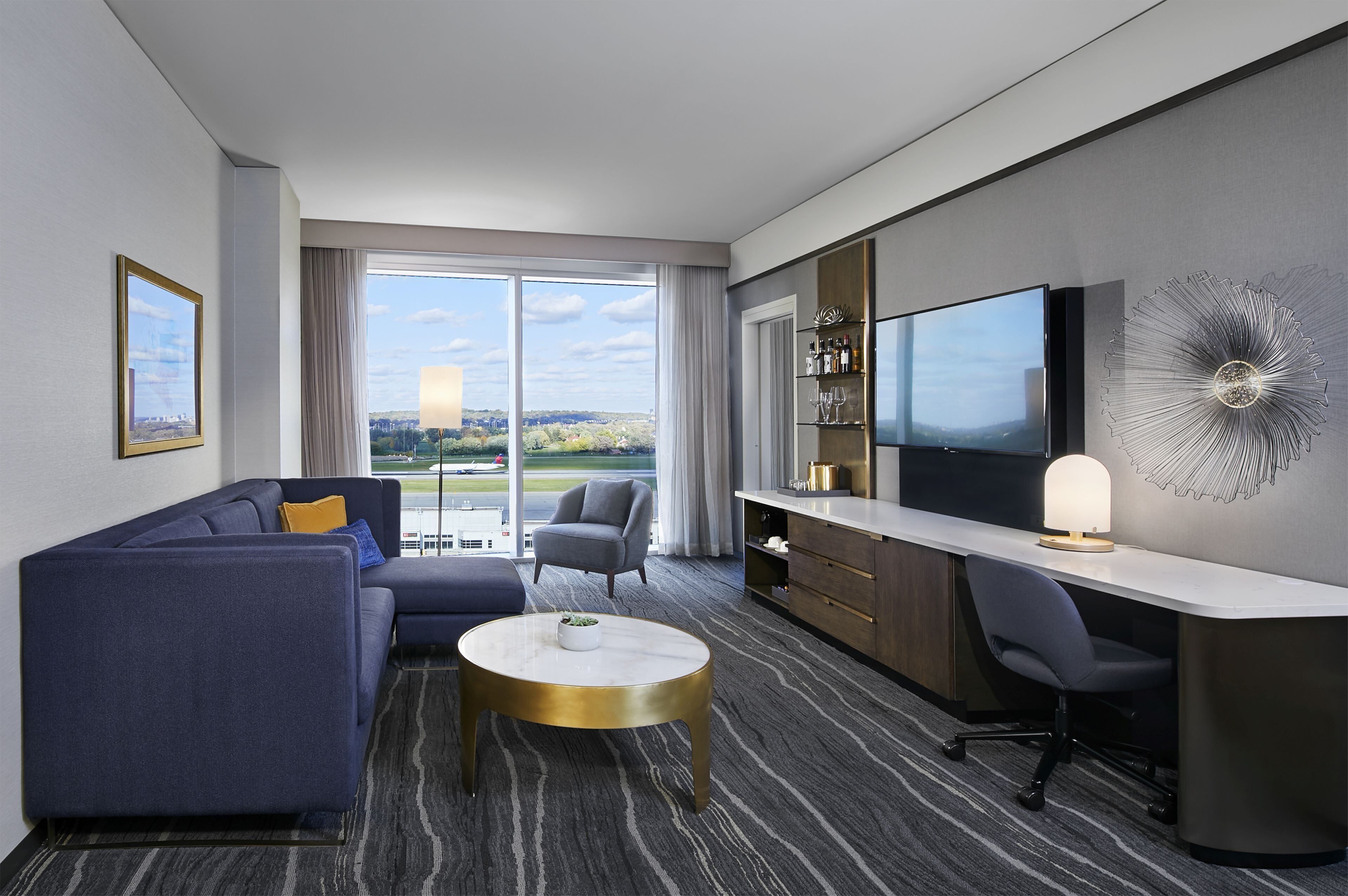 Una habitación de hotel moderna con sofá, sillón, escritorio y un televisor grande.
