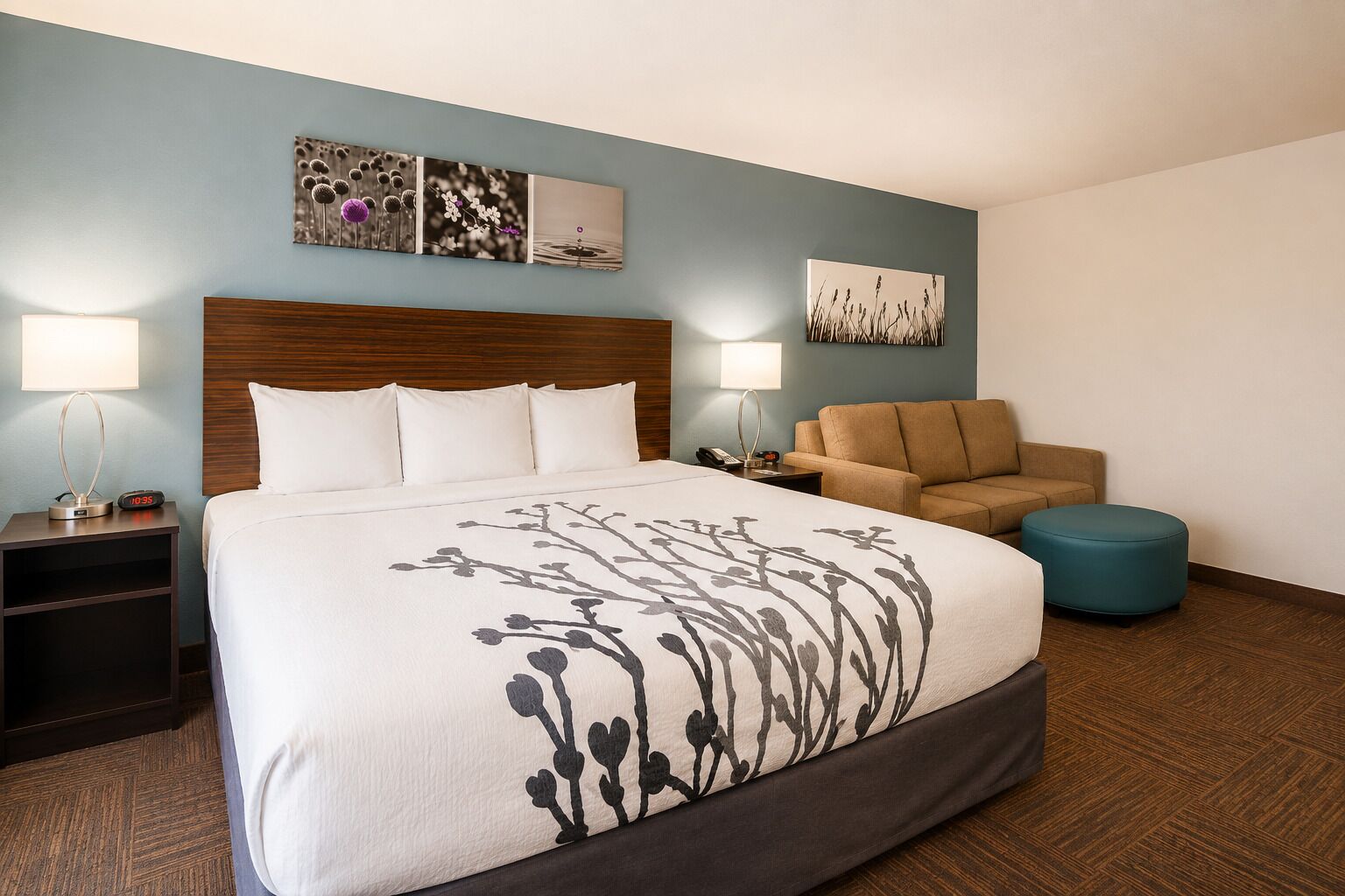 A hotel room with a large bed, two bedside tables, a sofa, a round ottoman, and wall art.