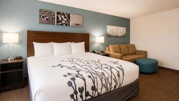 A hotel room with a large bed, two bedside tables, a sofa, a round ottoman, and wall art.