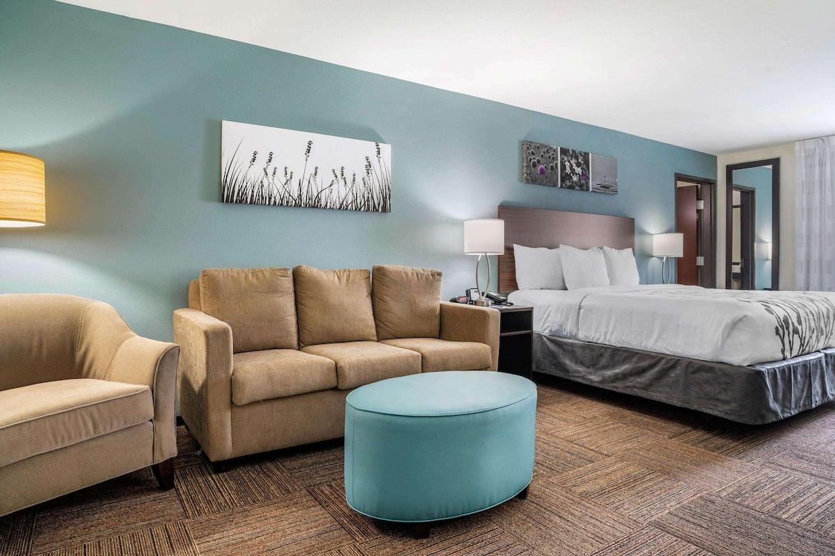 A hotel room with a bed, two sofas, a round ottoman, and a nightstand with a lamp.