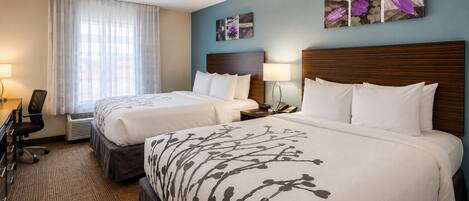 A hotel room with two beds, a desk, and a chair. There are wall-mounted lamps and artwork.