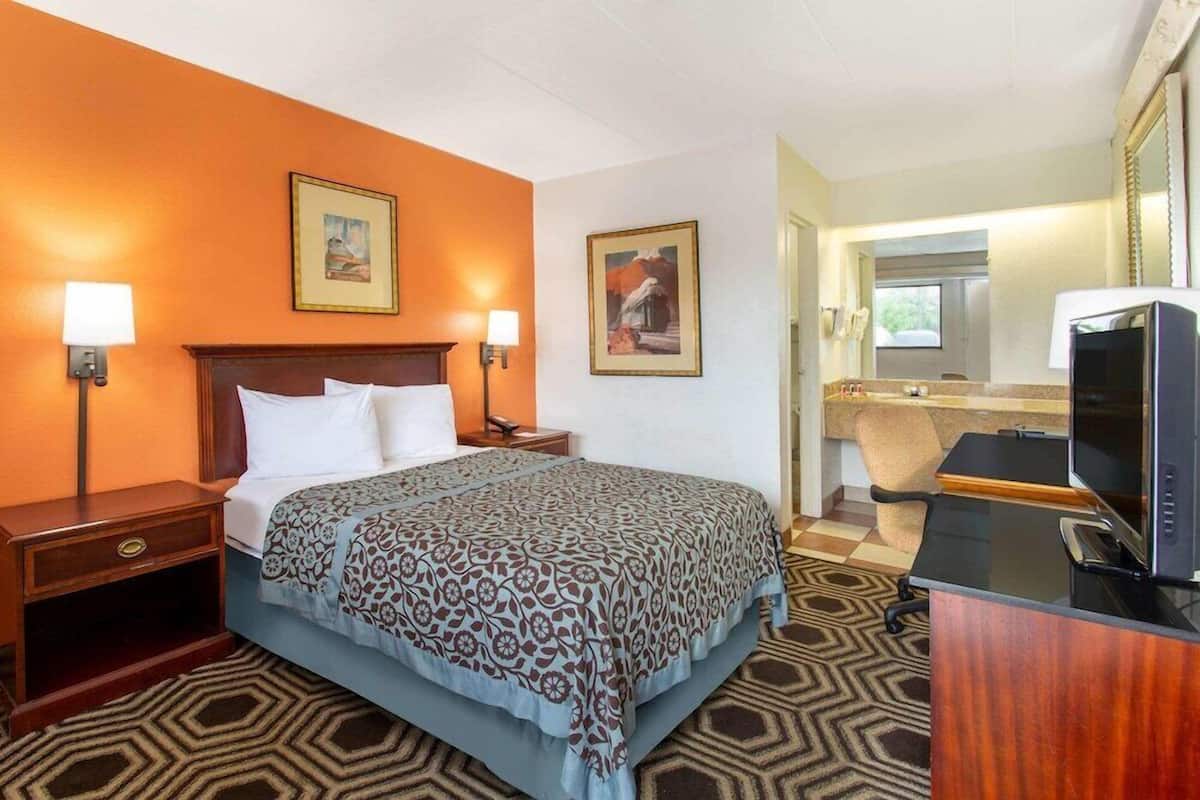 Habitación de hotel con cama, lámparas de noche, escritorio con televisor, baño con lavamanos y espejo, y dos cuadros en la pared.
