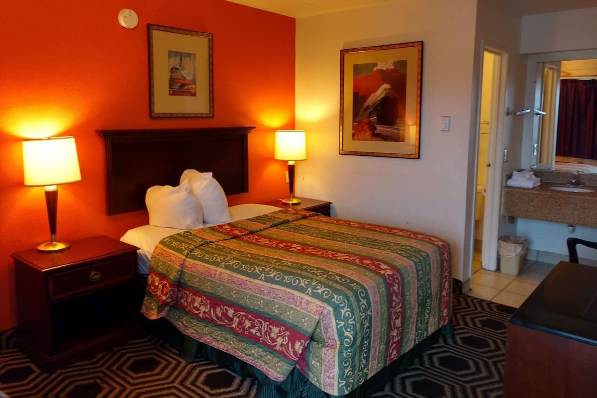Una habitación de hotel con una cama, dos lámparas de noche, un cabecero, un cuadro en la pared y un baño a la vista a través de una puerta abierta.