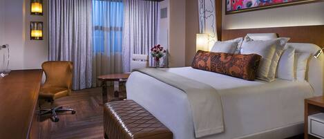 A hotel room with a large bed, a desk with a chair, a small table, and a bench.