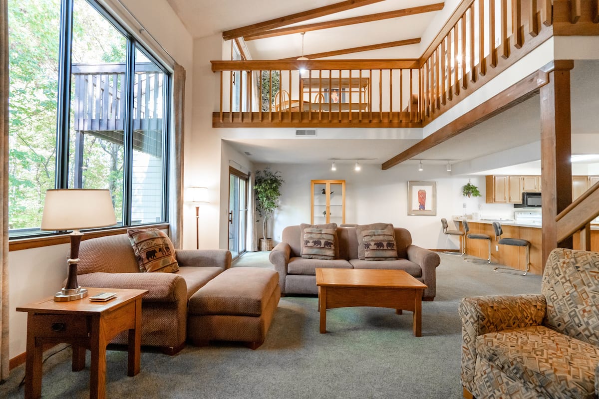 A spacious living room with a high ceiling, a wooden staircase, and a view of the outdoors through large windows.