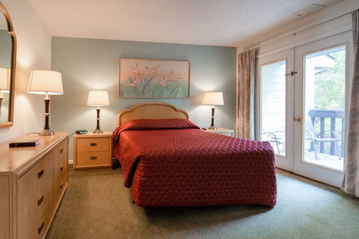 A bedroom with a bed, bedside lamps, a nightstand, a dresser, and a large window with a view of the outdoors.