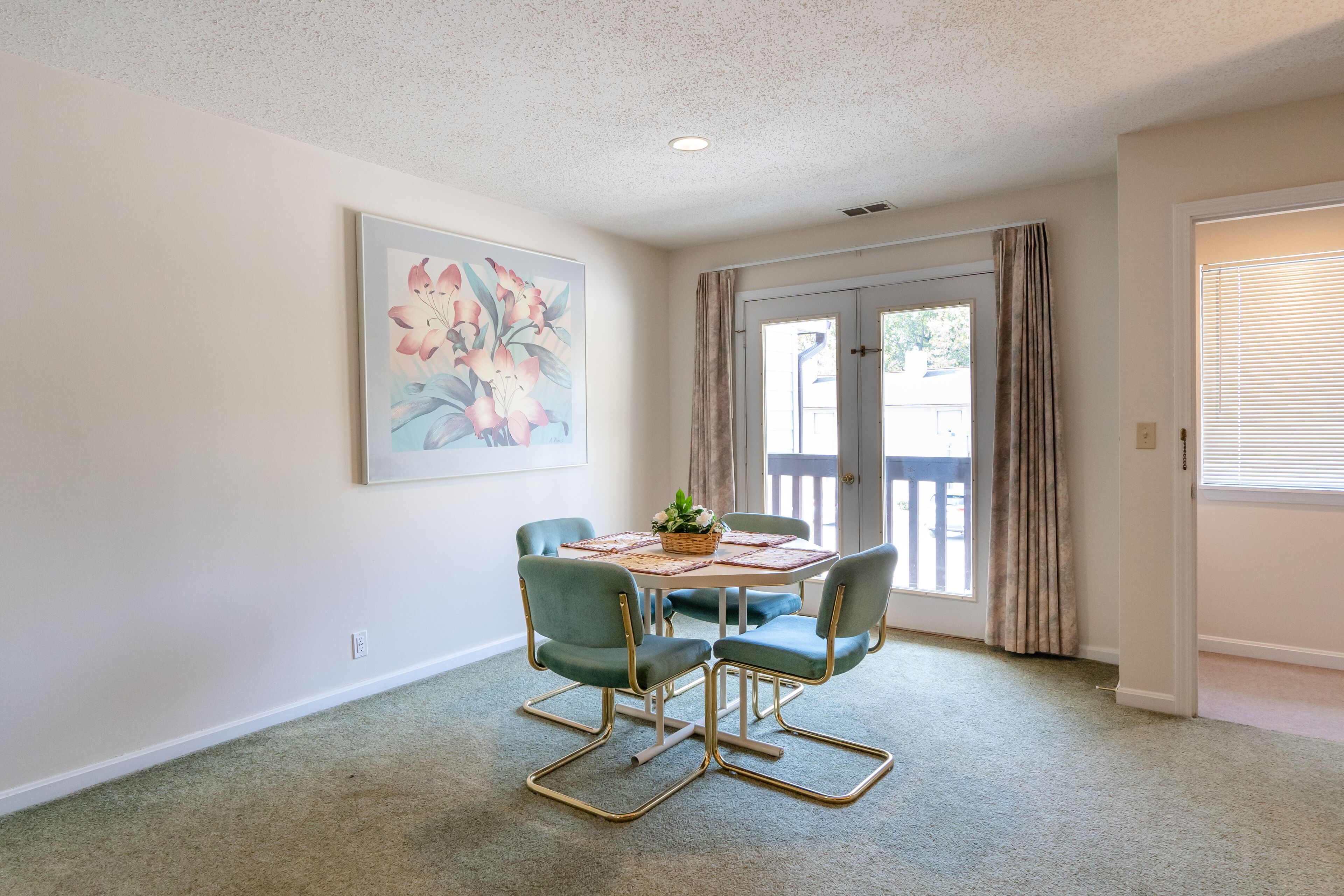 A dining area with a table and four chairs, a painting on the wall, and a door leading outside.
