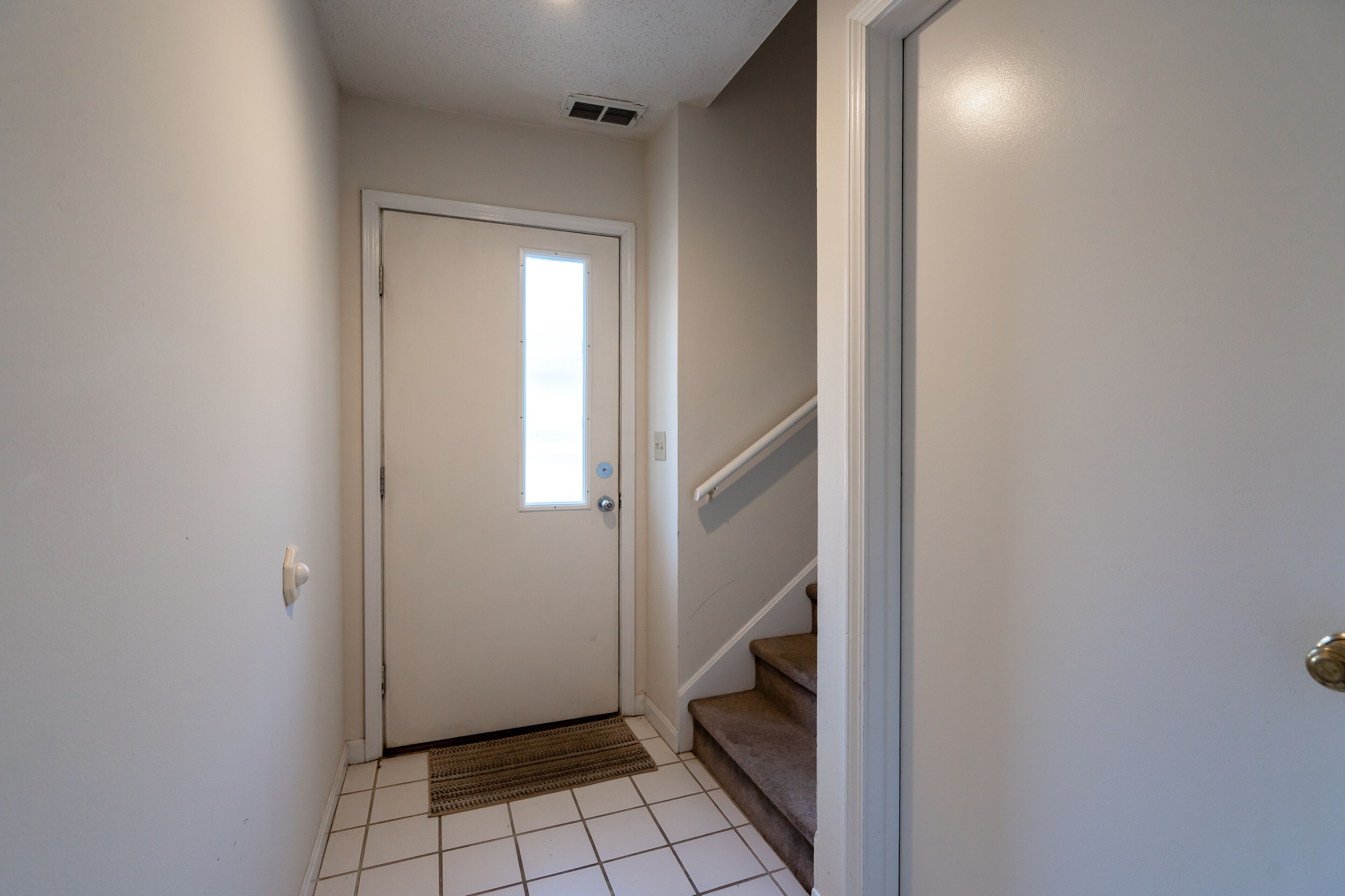 Un couloir avec une porte blanche, une petite fenêtre et un escalier recouvert de moquette.