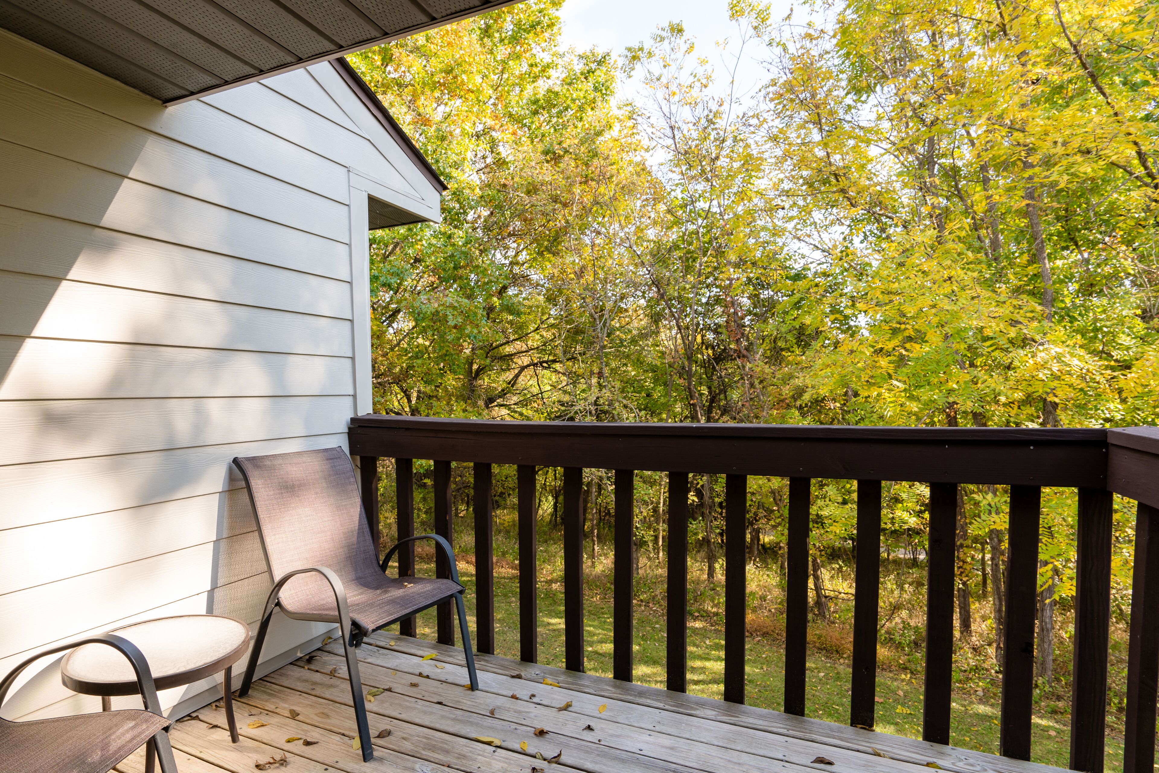 A wooden deck with two chairs, a white building, and a forested area in the background.