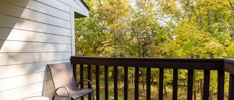 A wooden deck with two chairs, a white building, and a forested area in the background.