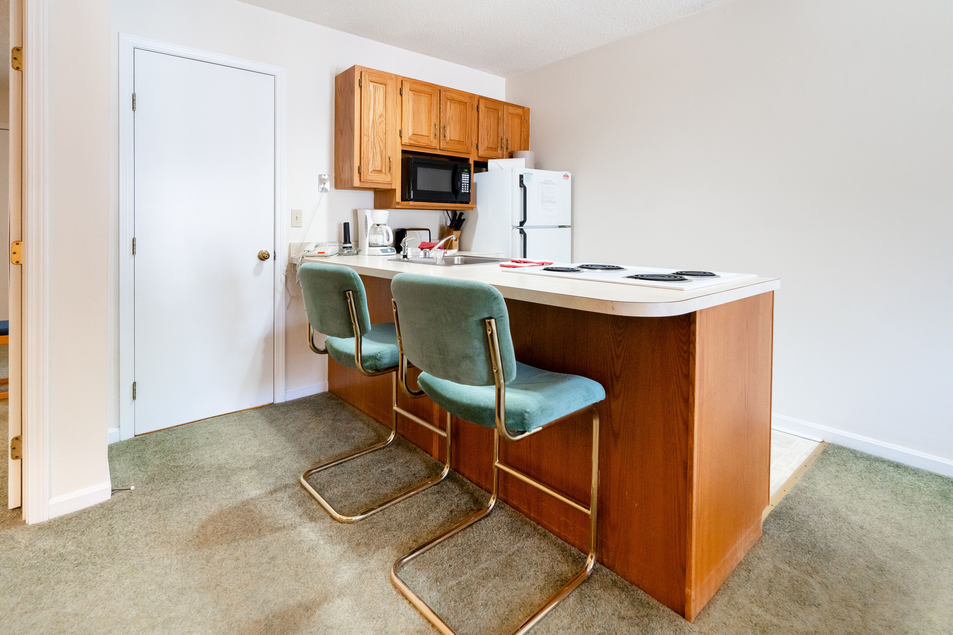 A compact kitchen with wooden cabinets, a microwave, a refrigerator, and two bar stools.