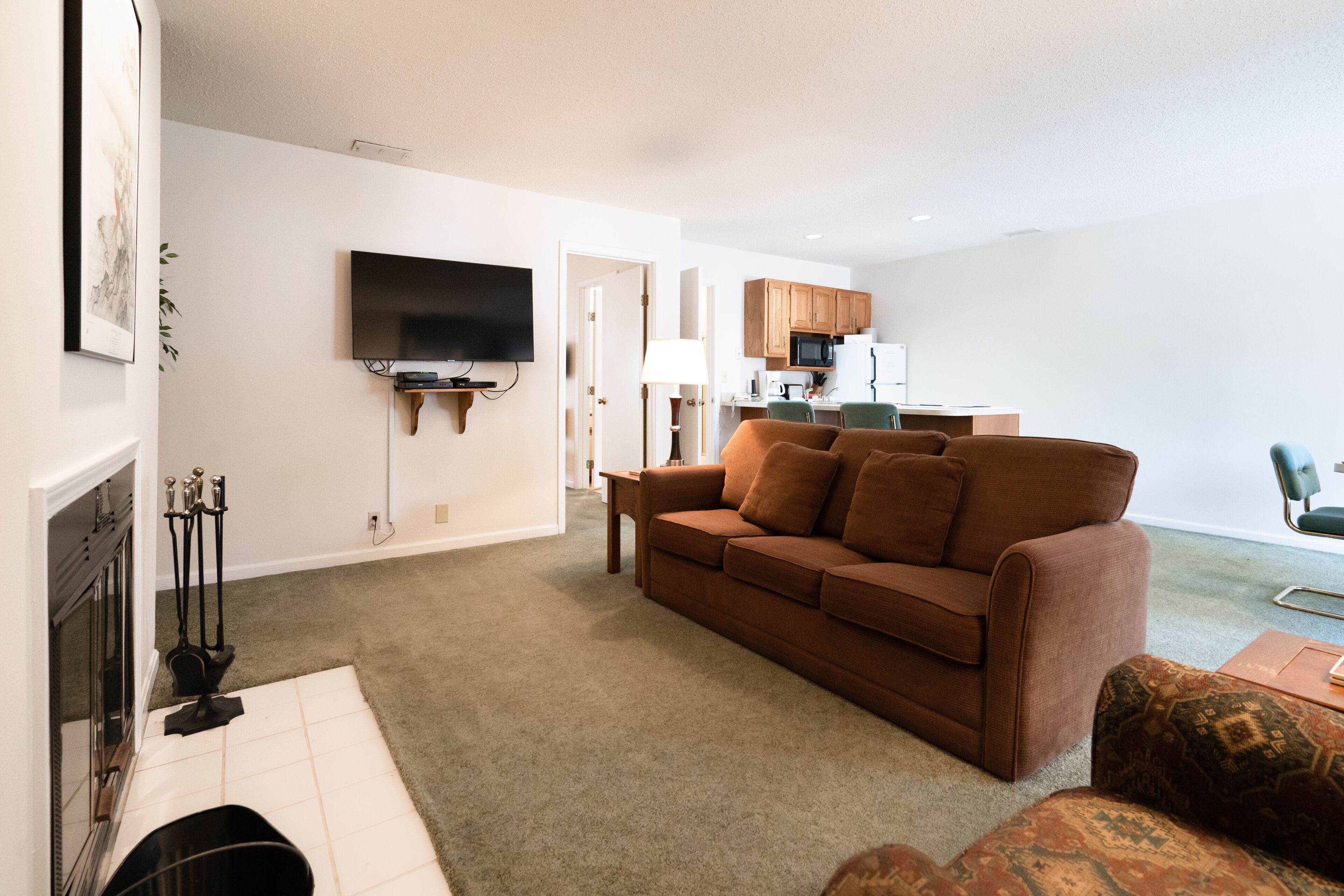 A living room with a brown sofa, a television mounted on the wall, a fireplace, and a kitchen area in the background.