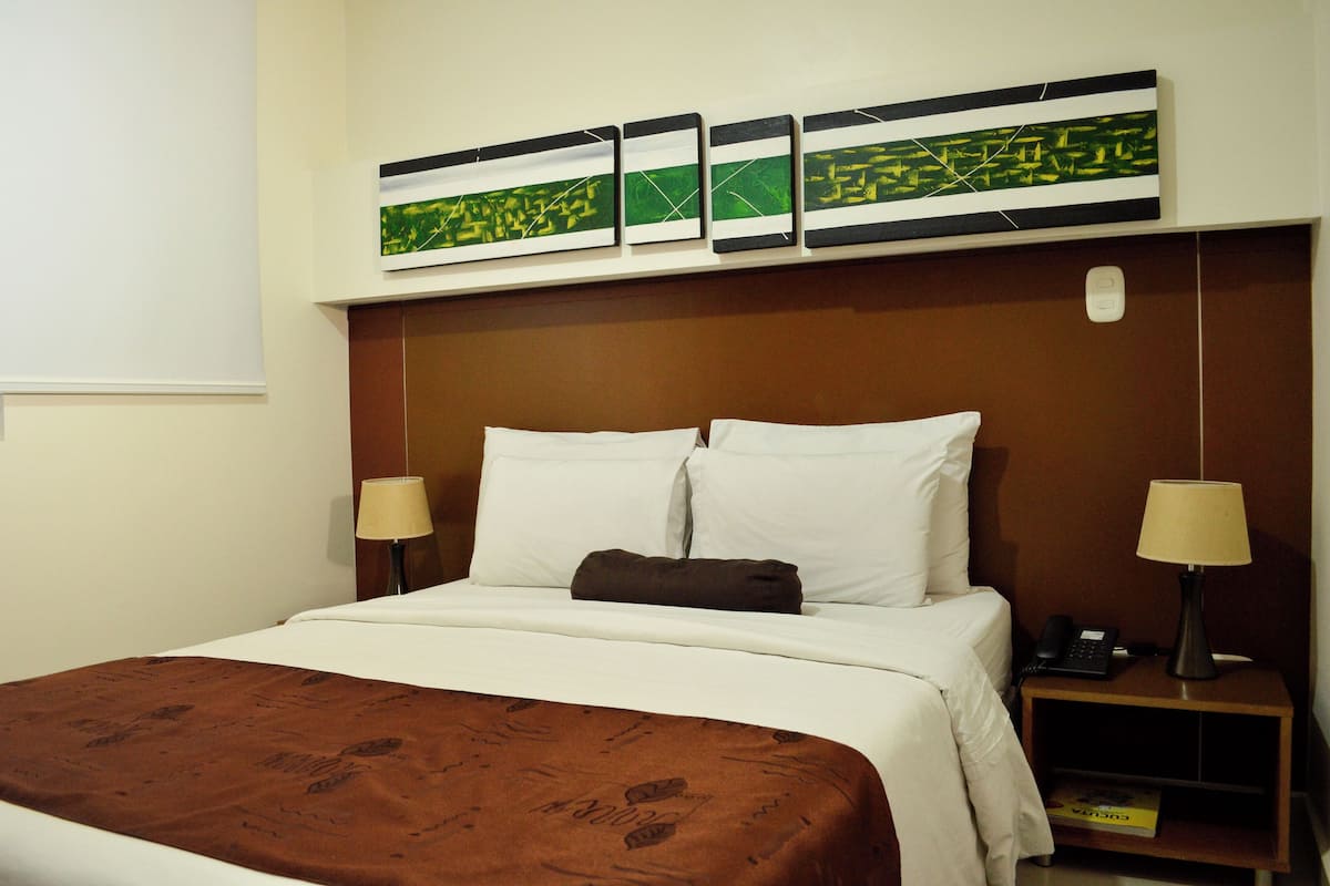 Een hotelkamer met een groot bed, twee bedlampen, een nachtkastje met een telefoon en een boek, en drie ingelijste kunstwerken aan de muur.
