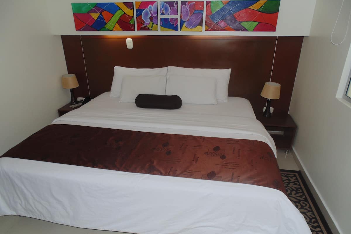 Hotelkamer met een groot bed, wit beddengoed, een bruine plaid en een hoofdeinde met een lichtschakelaar. Twee nachtlampjes en een raam met uitzicht.