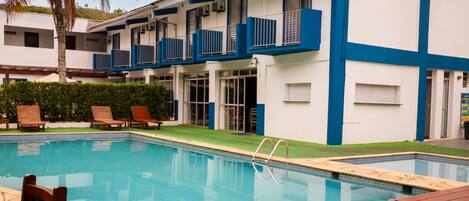 Uma piscina com deck de madeira, espreguiçadeiras e um edifício com varandas.