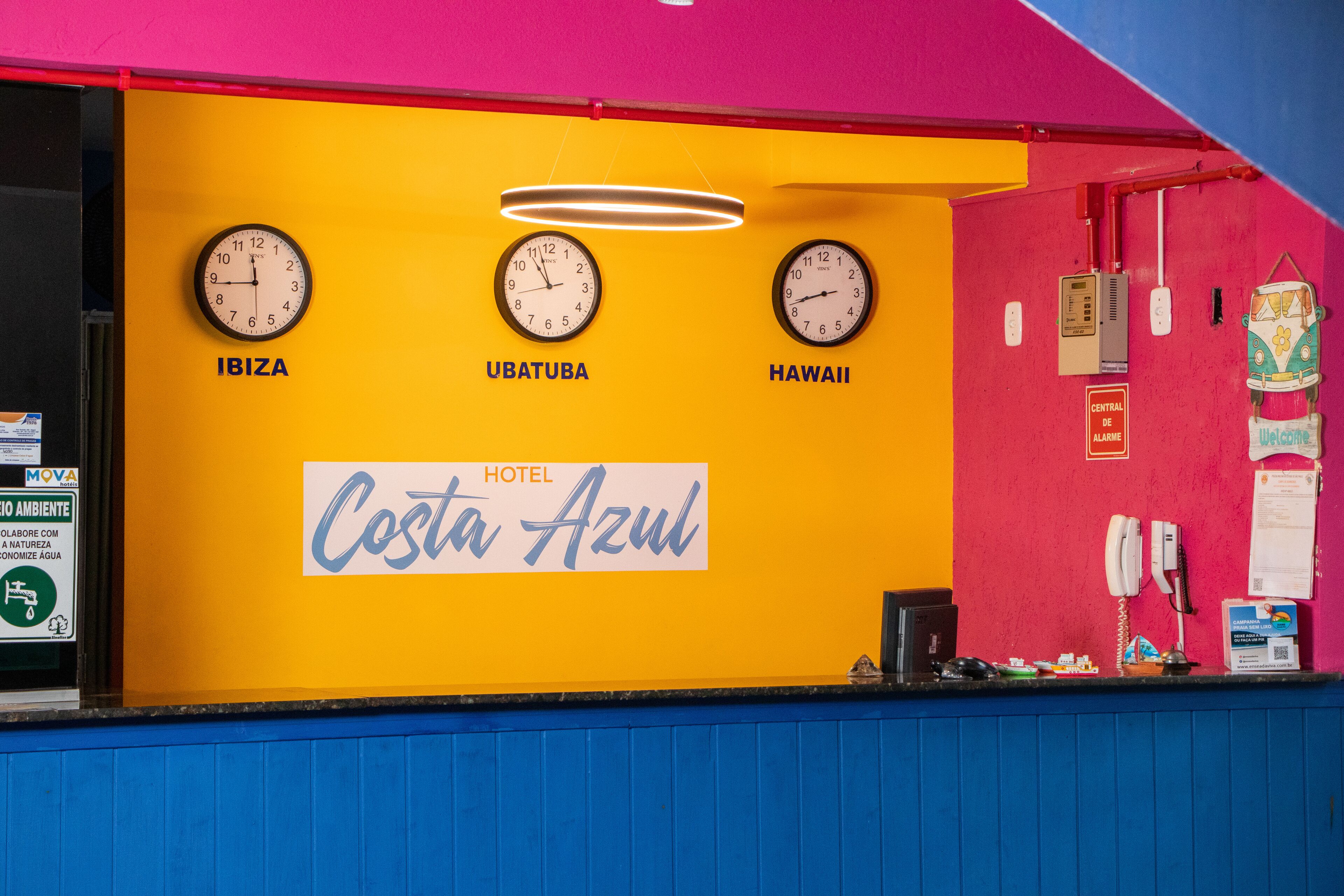 A hotel reception desk with clocks labeled 'IBIZA,' 'UBATUBA,' and 'HAWAII,' and a sign reading 'HOTEL Costa Azul.'